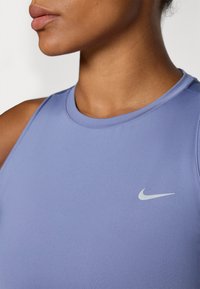 Fioletowy top sportowy Nike z okrągłym dekoltem i bezrękawnikowym krojem. Posiada małe, białe logo Nike na lewej stronie klatki piersiowej.