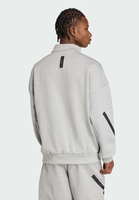 Svetlo šedá mikina s prehnaným strihom, rebrovaným lemom a čiernymi akcentmi na rukávoch. Má detail s nášivkou na chrbte. Mäkká textúra.