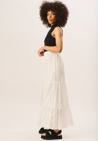 Black sleeveless top, white eyelet maxi skirt with tiered ruffles and floral cutouts. Black chunky sandals with straps.