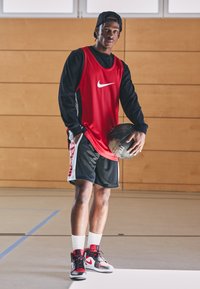 Rød Nike basketballtrøye med hvit swoosh, svarte shorts og svarte, røde og grå sneakers. Holder en svart basketball.