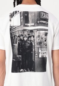 White t-shirt featuring a black and white graphic of a group standing by a London bus. Design includes detailed faces and vintage bus elements.