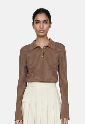 Donna con capelli scuri raccolti indietro indossa un maglione polo marrone a costine a maniche lunghe e una gonna plissettata color crema a vita alta su uno sfondo semplice.
