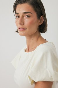 Witte jurk met pofmouwen, een ronde halslijn en een gestructureerde stof. Natuurlijke make-up en kort haar, tegen een neutrale achtergrond.
