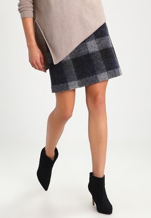 Femme portant un haut beige à manches longues, une jupe à carreaux gris et noir, et des bottines en daim noires à bout pointu et talons hauts.