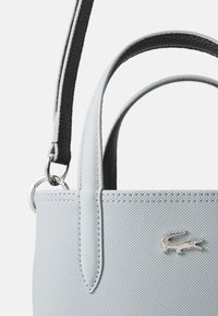Sac à main synthétique gris avec une finition texturée, des poignées rondes et une bandoulière noire contrastante. Présente un détail en logo crocodile argenté.