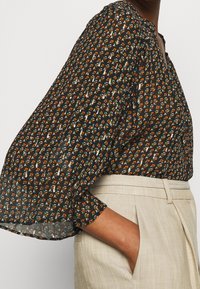 Svart blommig blus med utsvängda ärmar, prydd med små mönster i orange och turkos, bärs inskjuten i beige högmidjade byxor.