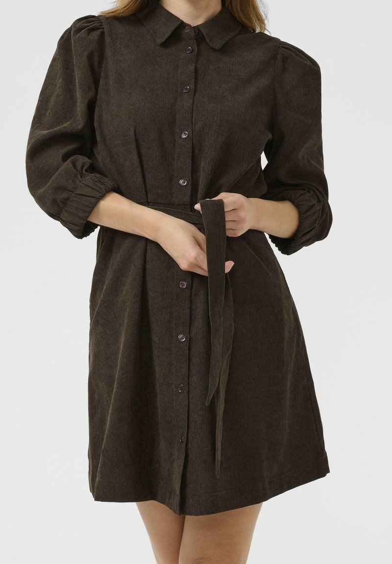 Femme portant une robe boutonnée marron foncé avec des manches bouffantes, nouée à la taille avec une ceinture en tissu assortie, sur un fond uni.
