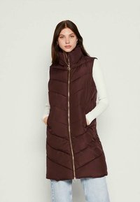 Colete puffer castanho escuro com gola alta, fecho frontal, bolsos laterais e padrão de costura em zigue-zague. Tecido leve com uma textura suave.