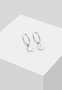 Boucles d'oreilles créoles argentées avec breloques étoile et croissant de lune sur une surface géométrique blanche.