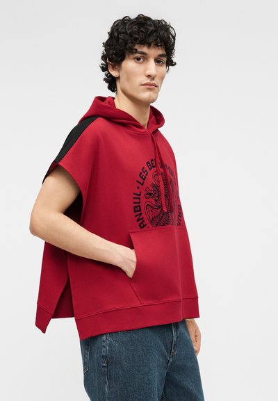 Jonge man met krullend haar, gekleed in een rood mouwloos vest met een zwarte opdruk en een blauwe spijkerbroek, staand met één hand in zijn zak.