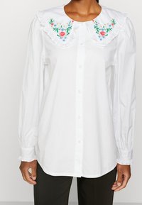 Camicia di cotone bianca con colletto arricciato, bordata da colorati ricami floreali e chiusura a bottoni. Le maniche lunghe hanno polsini elastici.