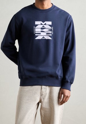 Mand iført mørkeblå sweatshirt med fedt hvidt geometrisk tekstdesign på brystet, kombineret med lyse beige bukser, neutral baggrund.