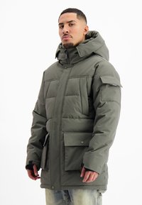 Man in een olijfgroene gewatteerde winterjas met capuchon, grote frontzakken en klittenband manchet, staande tegen een witte achtergrond.