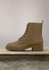 Bottine en daim beige à lacets avec talon épais et languette de tirage sur une surface en bois sur fond neutre.