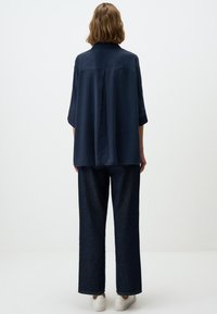 Marineblaue Kurzarmbluse mit geradem Schnitt und Rückenfalte, kombiniert mit weit geschnittenen dunkelblauen Jeans und weißen Sneakers.
