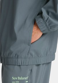 Persona vestida con chaqueta y pantalones grises de New Balance, con la mano en el bolsillo de la chaqueta, mostrando detalles de los puños y la cinturilla elásticos.