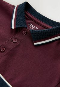 Burgunderfarvet polo med en tekstureret navykrave, tre knapper og hvide-røde striber på kraven. Labelen står "NEXT."
