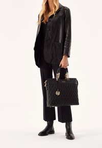 Czarna pikowana torba na ramię z złotymi akcentami, podwójnymi uchwytami u góry oraz zawieszką z logo, zestawiona z czarną skórzaną kurtką i spodniami.