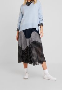 Pull en tricot bleu clair avec un col en V, superposé à une jupe plissée noire et marine présentant des motifs variés et des rayures blanches, assortie à des baskets blanches.