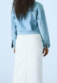 Persona con capelli ricci che indossa una giacca di denim blu chiaro e una gonna bianca a vita alta, in piedi contro uno sfondo blu pallido.