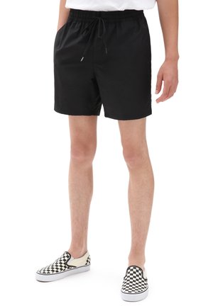 Persona che indossa pantaloncini neri con coulisse e scarpe slip-on a quadri bianche e nere, in piedi con una mano rilassata lungo il corpo.