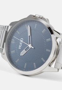 Montre argentée avec un cadran bleu, présentant des marqueurs d'heure, des accents métalliques et un bracelet en acier inoxydable. Le nom de la marque "HUGO" apparaît sur le cadran.