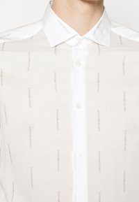 Biała koszula z guzikami z delikatnym beżowym wzorem z logo marki "Emporio Armani". Kołnierzyk oraz listwa na guziki są gładkie, białe.