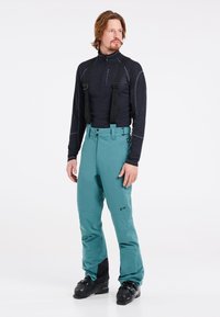 Pantalones de esquí en color aguamarina con tirantes negros ajustables, puños negros reforzados y un diseño impermeable. Llevados con una camiseta negra de manga larga.