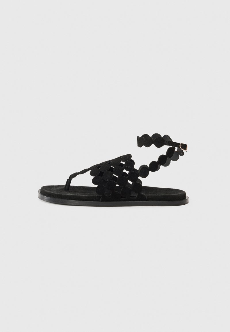 Sort sandal med vævet design lavet af ruskind; har flad sål, ankelrem med afrundede detaljer og minimal hardware.