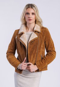 Model trägt eine braune Wildlederjacke mit einem Lammfellkragen und posiert mit den Händen in der Nähe des Reißverschlusses der Jacke, vor einem einfarbigen Hintergrund.