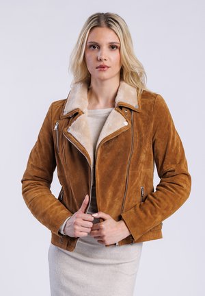 Model in een bruine suède jas met een schapenvachtkraag, poserend met haar handen nabij de rits van de jas, tegen een effen achtergrond.