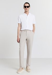 Chemise blanche à manches courtes avec boutons, pantalon beige à rayures fines et chaussures légères à enfiler. Des lunettes de soleil sombres complètent le look.