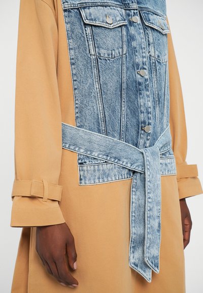 Manteau en tissu beige avec un panneau avant en denim bleu et une taille à nouer. Dispose de poches poitrine à boutons et de manches roulées avec des accents de boucle.