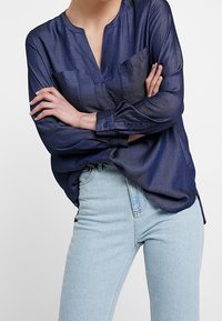 Blusa trasparente blu navy con scollatura a V, maniche lunghe e due tasche sul petto. Indossata infilata dentro a jeans azzurri.