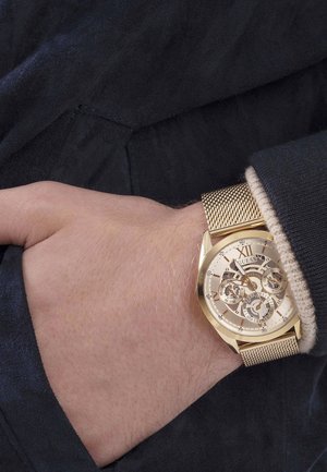 Gouden metalen horloge met een mesh band. Heeft een skeletwijzerplaat, Romeins cijfer XII en gedetailleerde subwijzerplaten. Liggend op een gestructureerde mouw.