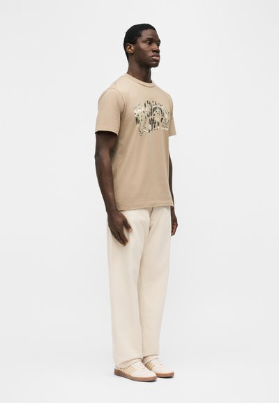 Άνδρας στέκεται στραμμένος προς τα δεξιά, φοράει μπεζ μπλουζάκι με στάμπα, φαρδιά κρέμ παντελόνια και ανοιχτό μπεζ αθλητικά παπούτσια, με φόντο απλό λευκό τοίχο.