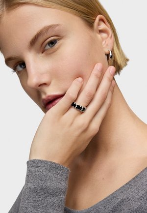 Anello d'argento con motivi neri a trifoglio, finitura liscia. Modella che indossa un maglione grigio e piccoli orecchini a cerchio d'argento, mano appoggiata sul volto.
