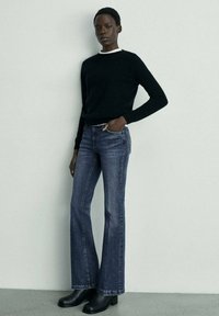 Černý svetr, modré zvonové džínsy s klasickým prošitím a černé kotníkové boty. Hladká textura s přiléhavým siluetu. Neutrální pozadí.