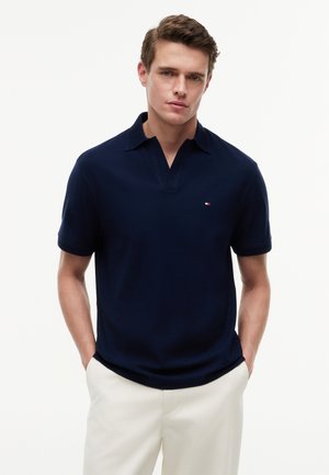 Młody mężczyzna w granatowej koszulce polo z krótkim rękawem z małym logo i białych spodniach, ręce w kieszeniach, neutralne tło.