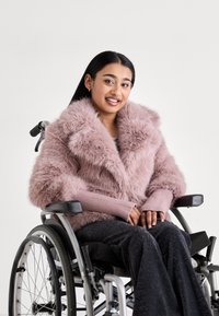Jeune femme souriante en fauteuil roulant portant un manteau en fausse fourrure rose et un pantalon foncé, sur un fond clair uni.
