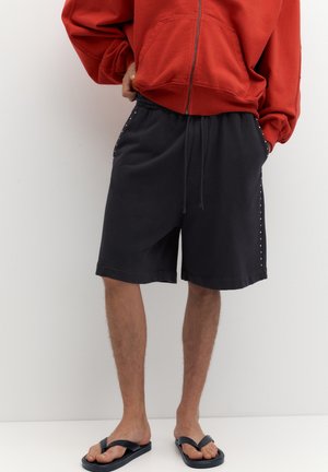 Person trägt einen roten Hoodie mit Reißverschluss, schwarze Shorts mit Kordelzug und seitlichen Nieten sowie schwarze Flip-Flops und steht vor einem weißen Hintergrund.
