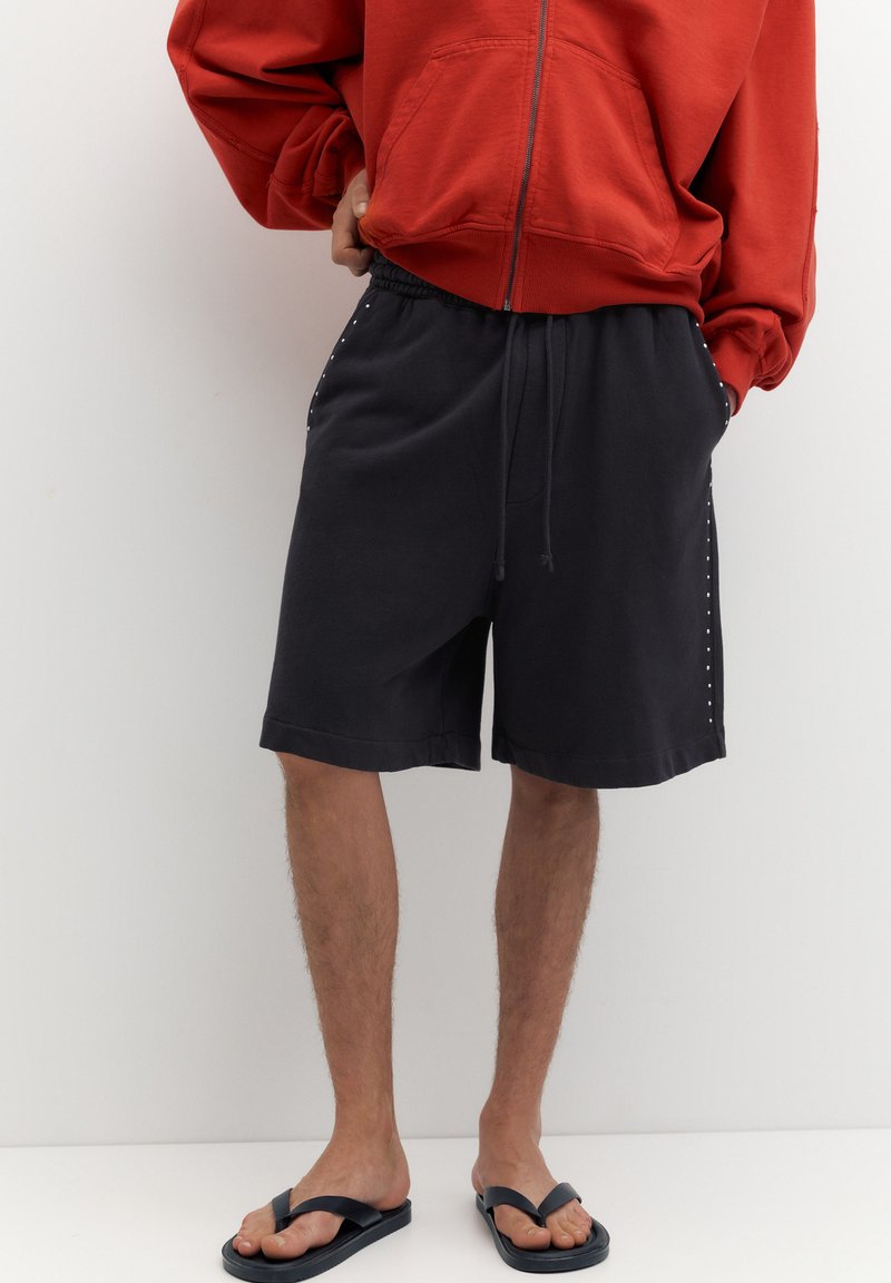 Personne portant un sweat à capuche rouge zippé, un short noir à cordon avec clous latéraux et des tongs noires, debout sur un fond blanc uni.