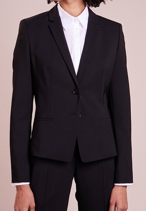 Žena nosí na míru šité černé sako se dvěma knoflíky přes bílou košili s límečkem, doplněné černými kalhotami.