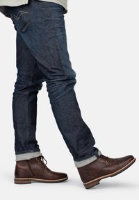 Braune Leder-Stiefeletten mit Schnürdesign und seitlichem Reißverschluss. Kombiniert mit dunklen Denim-Jeans, umgeschlagenen Säumen und dezenten Steppnähten.