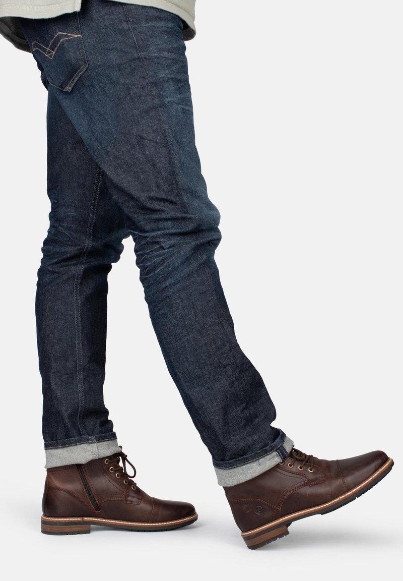 Braune Leder-Stiefeletten mit Schnürdesign und seitlichem Reißverschluss. Kombiniert mit dunklen Denim-Jeans, umgeschlagenen Säumen und dezenten Steppnähten.