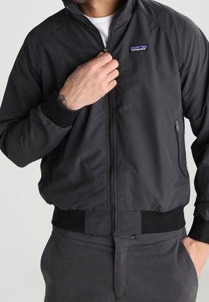 Mężczyzna noszący czarną kurtkę Patagonia, podciąga suwak do góry, z zapinaną na zamek boczną kieszenią oraz czarnymi prążkowanymi mankietami i pasem.