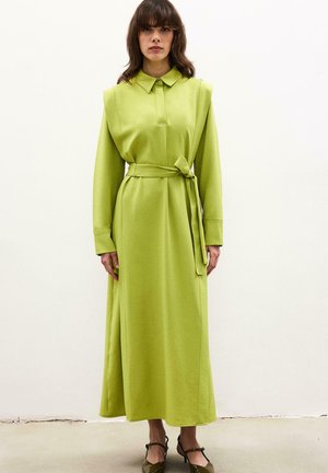 Kobieta w długiej limonkowo-zielonej sukience z poduszkami na ramionach, paskiem w talii i długimi rękawami, w połączeniu z ciemnymi butami na obcasie o spiczastym nosku.