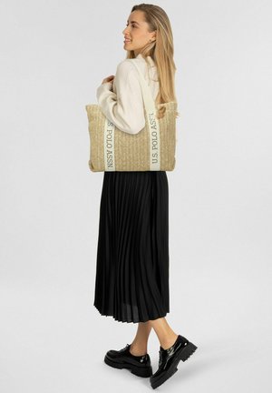 Vrouw in een beige trui en een zwarte geplooide rok draagt een grote geweven boodschappentas met de hengsels van "U.S. POLO ASSN.", en draagt zwarte plateauzolen.