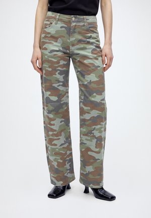 Personne portant un pantalon droit camouflage et des chaussures à talons noires, debout devant un fond clair uni.
