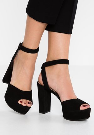 Sandalias de tacón alto de ante negro con suela de plataforma, diseño de punta abierta y correa ajustable en el tobillo con cierre de botón.